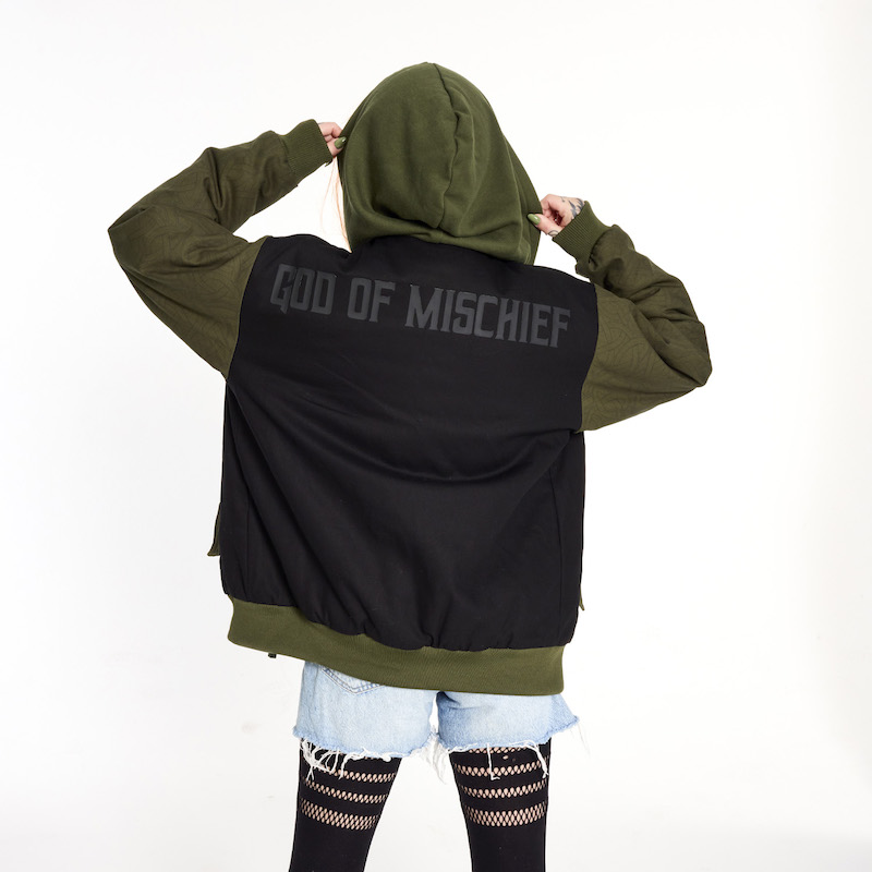 Woman facing away from camera and wearing the Loungefly COLLECTIV Marvel Loki The WEEKENDR Hooded Jacket, holding the green hood up to show off the words "God of Mischief" written on the back of the jacket. 